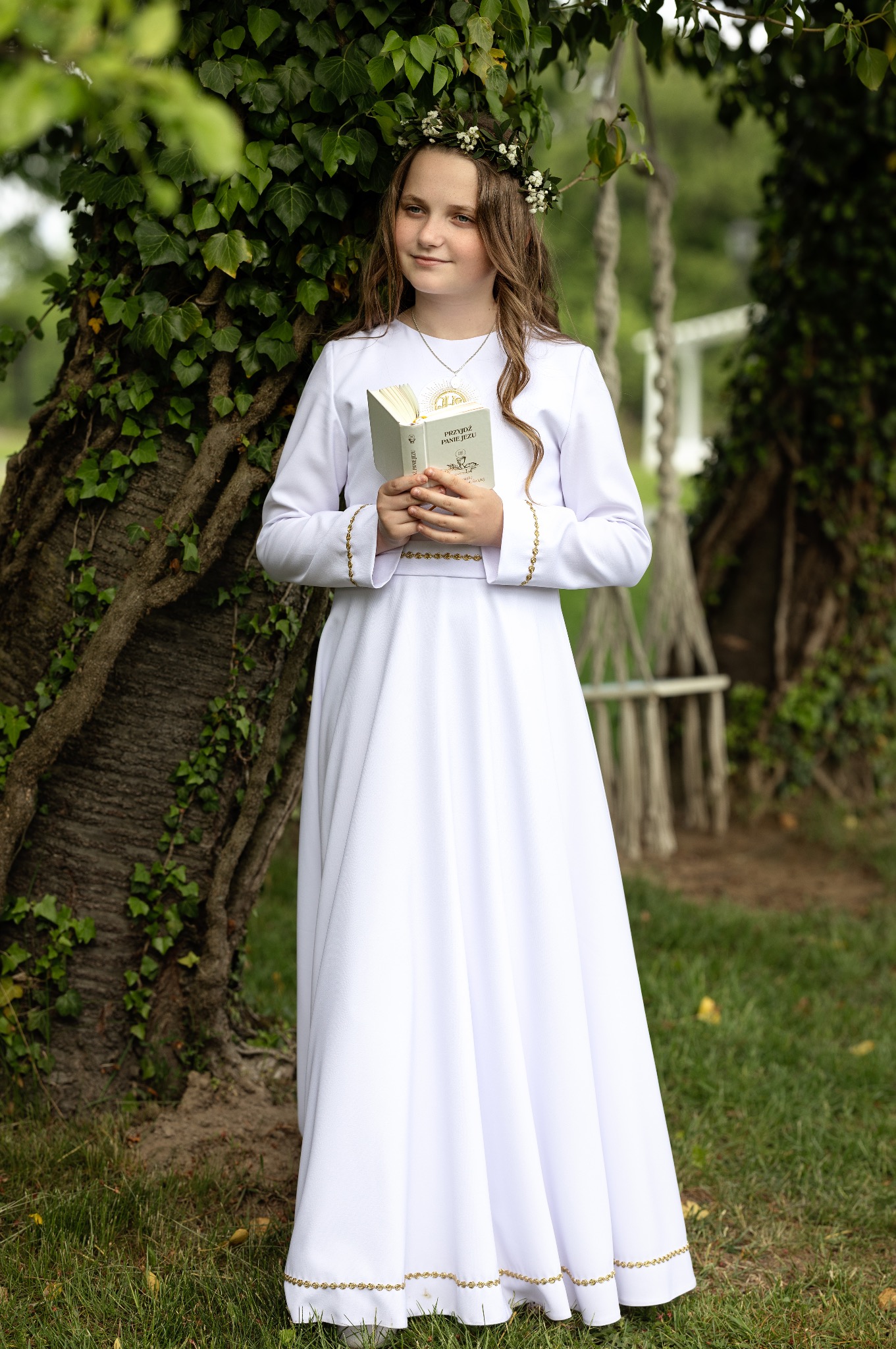Dziewczynka w białej sukience komunijnej z wiankiem na głowie, trzymająca książeczkę do Pierwszej Komunii Świętej, pozuje przy drzewie.