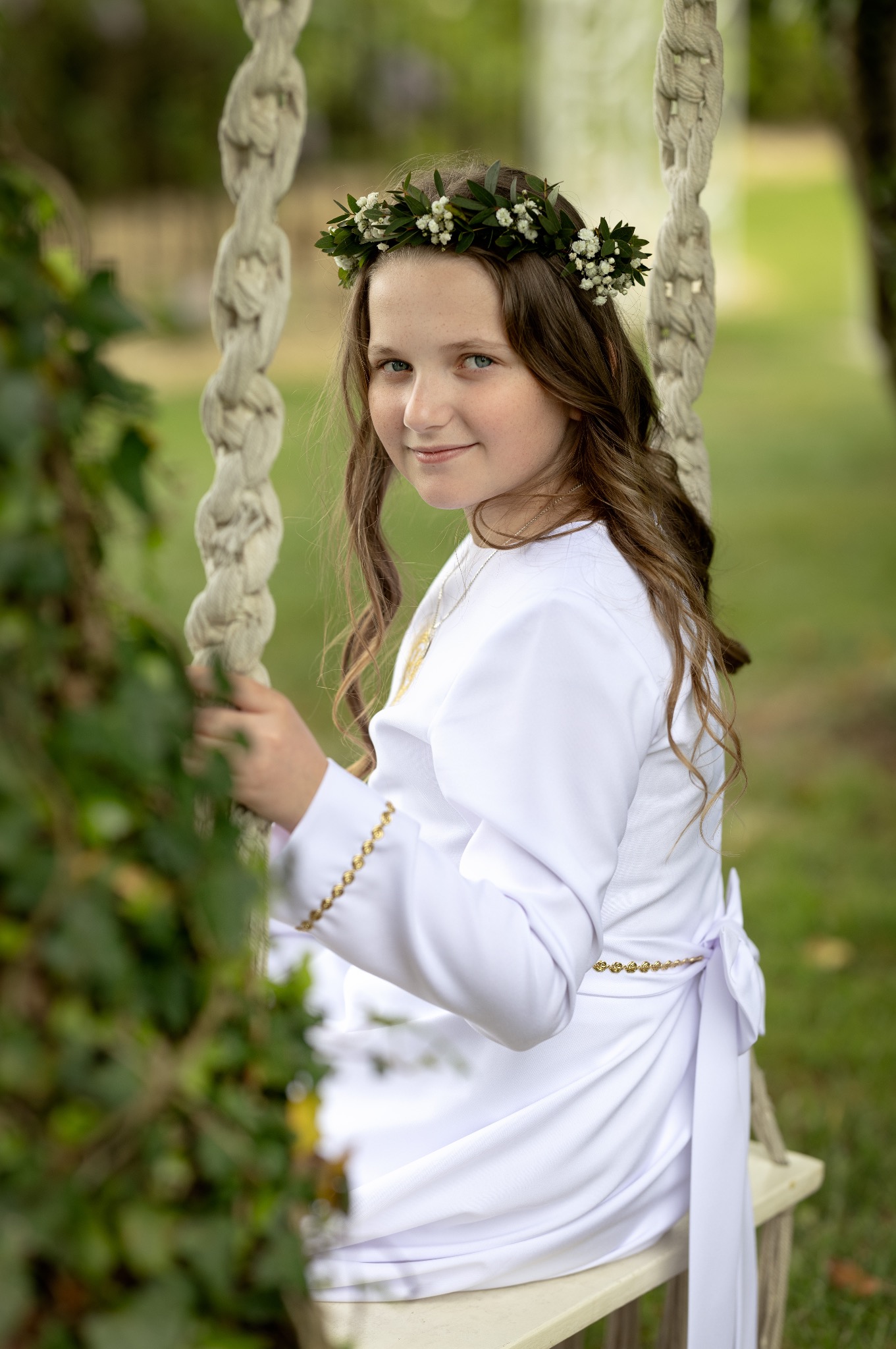 Dziewczynka w białej albie i wianku na głowie siedzi na huśtawce linowej w ogrodzie, spoglądając w obiektyw z delikatnym uśmiechem. Ujęcie portretowe, plener.