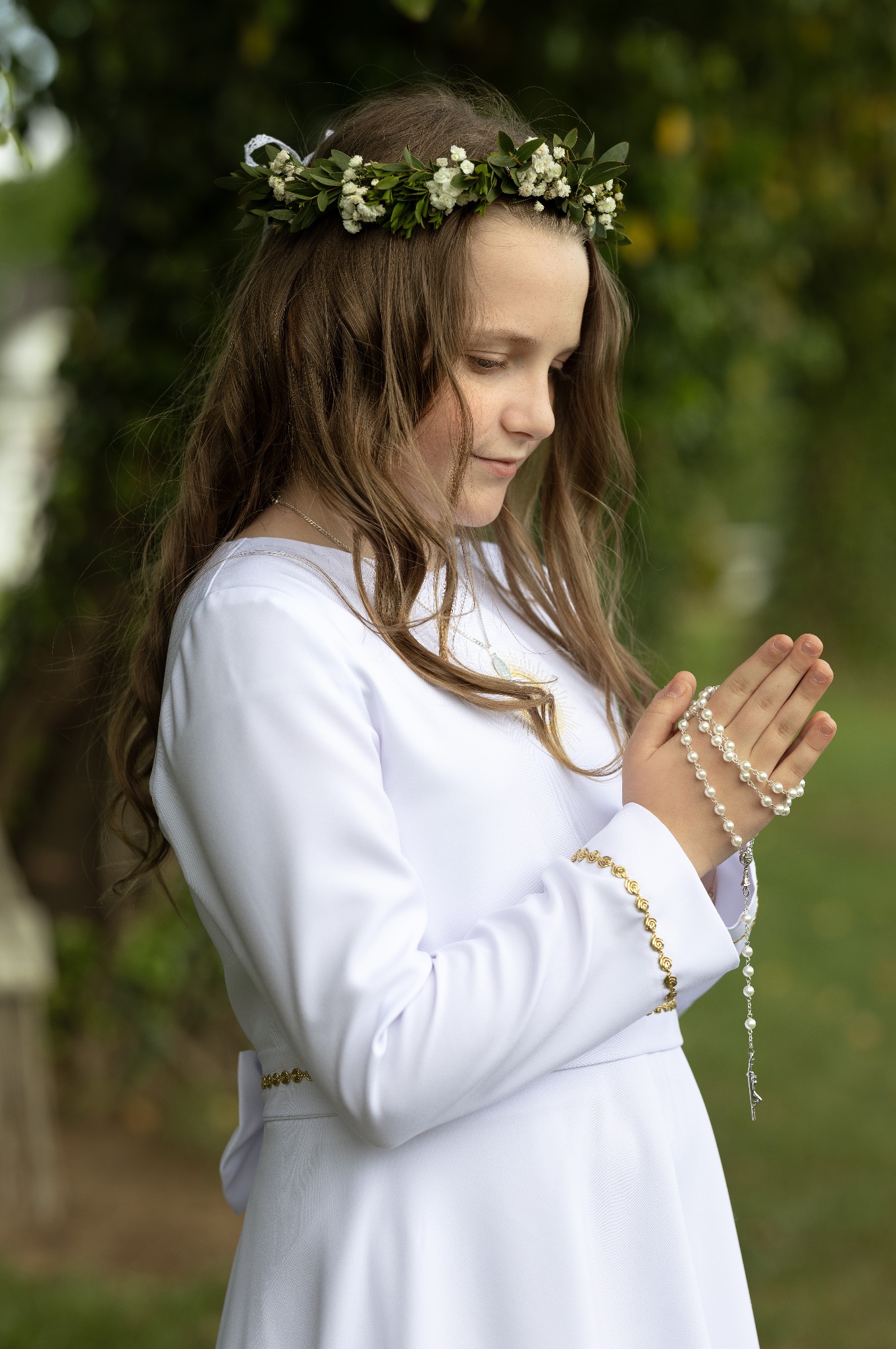 Dziewczynka w białej albie i wianku z kwiatów, z różańcem w dłoniach, spogląda w dół. Delikatne światło podkreśla detale stroju i skupienie na twarzy.