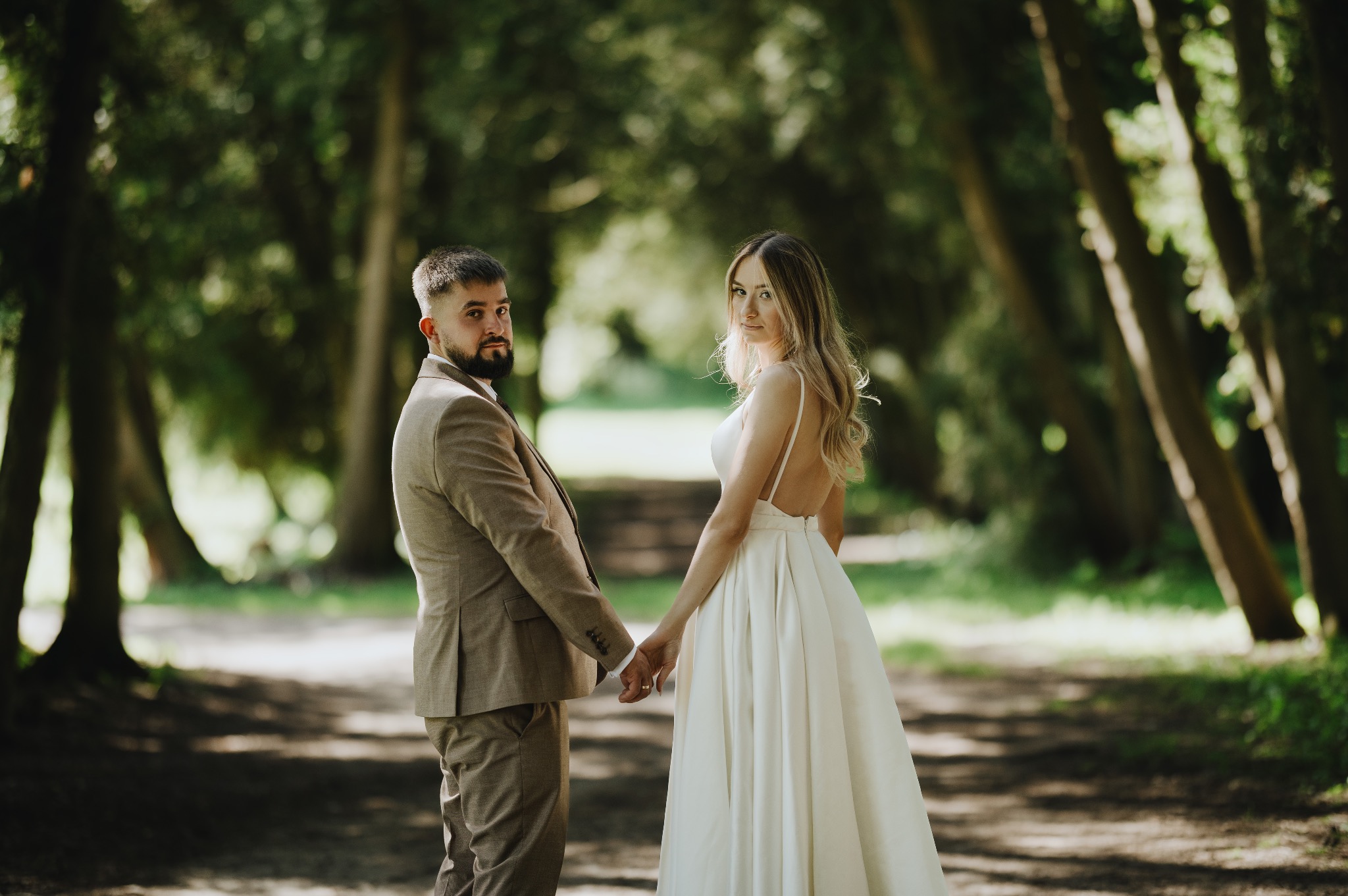 Para młoda w parku: mężczyzna w brązowym garniturze, kobieta w białej sukni z odkrytymi plecami, trzymają się za ręce, patrząc w obiektyw. Romantyczna sceneria z drzewami.