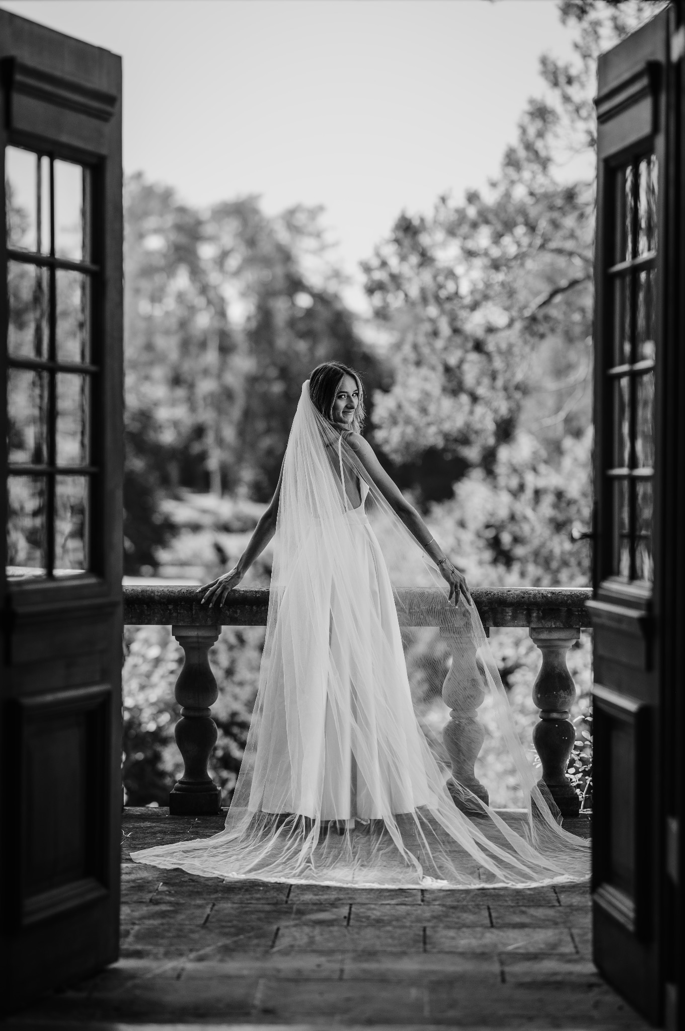 Czarno-białe zdjęcie kobiety w sukni ślubnej i welonie, stojącej na balkonie z otwartymi drzwiami, z widokiem na zieleń. Kobieta odwrócona, patrzy przez ramię.