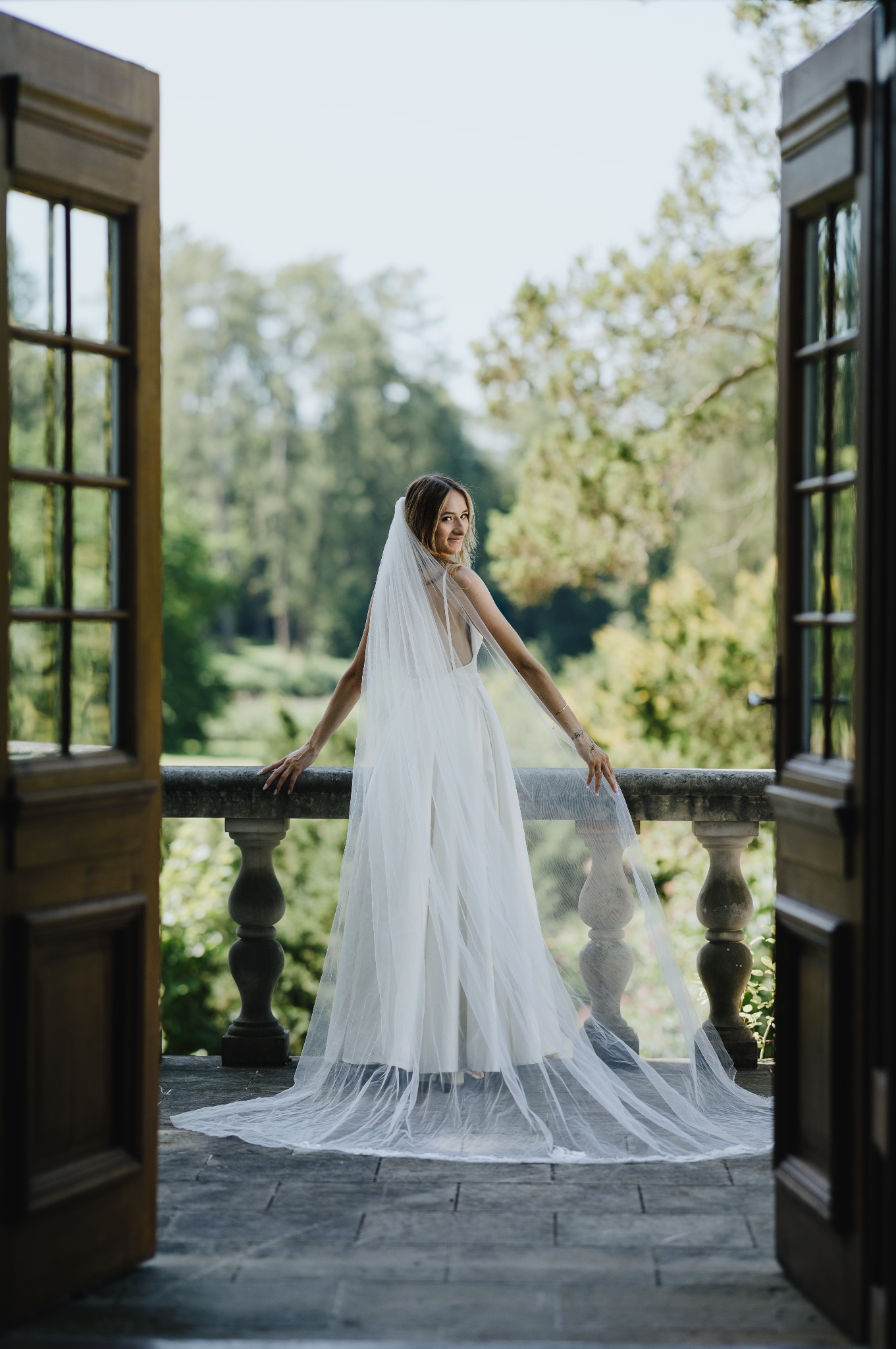 Panna młoda w białej sukni i welonie stoi na balkonie z kamienną balustradą, spoglądając przez ramię na tle zieleni. Ujęcie przez otwarte, drewniane drzwi.