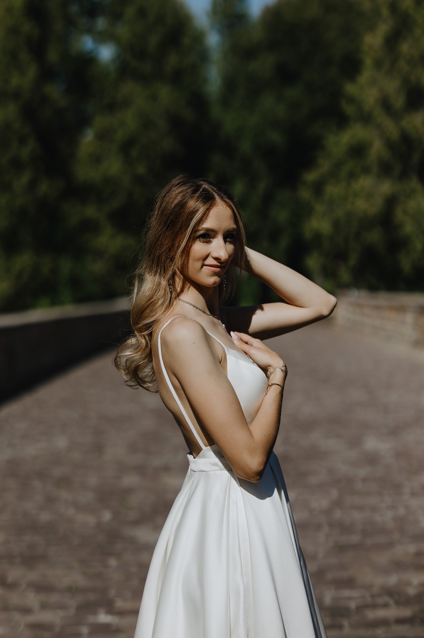 Portret kobiety w białej sukience na tle zieleni i kamiennego mostu. Ujęcie z boku, podkreślające delikatność i elegancję. Jasne, naturalne światło.