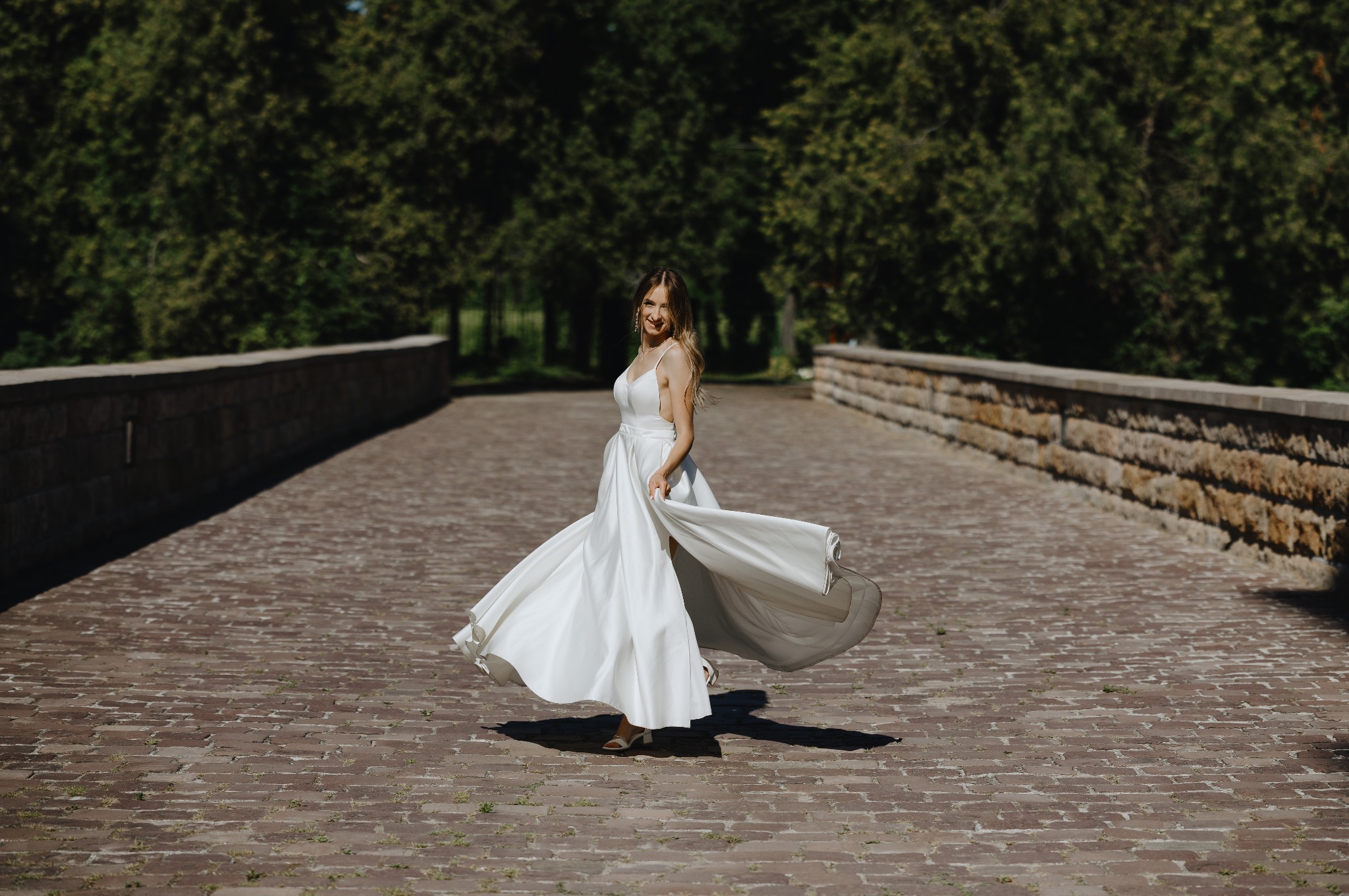 Uśmiechnięta panna młoda w białej sukni wiruje na kamiennym moście, otoczona zielenią drzew. Słoneczny dzień podkreśla blask sukni i radość chwili.
