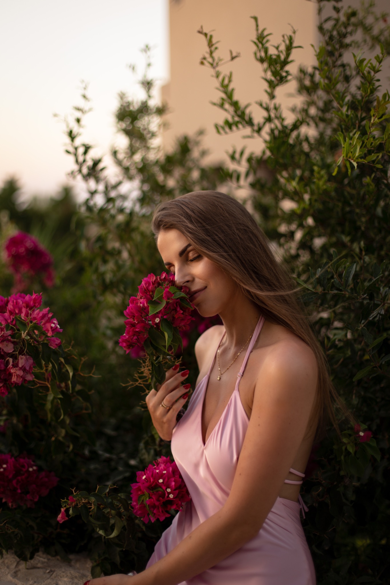 Kobieta w satynowej sukience wącha bukiet różowych kwiatów na tle zieleni i beżowego budynku w ciepłym świetle zachodzącego słońca.