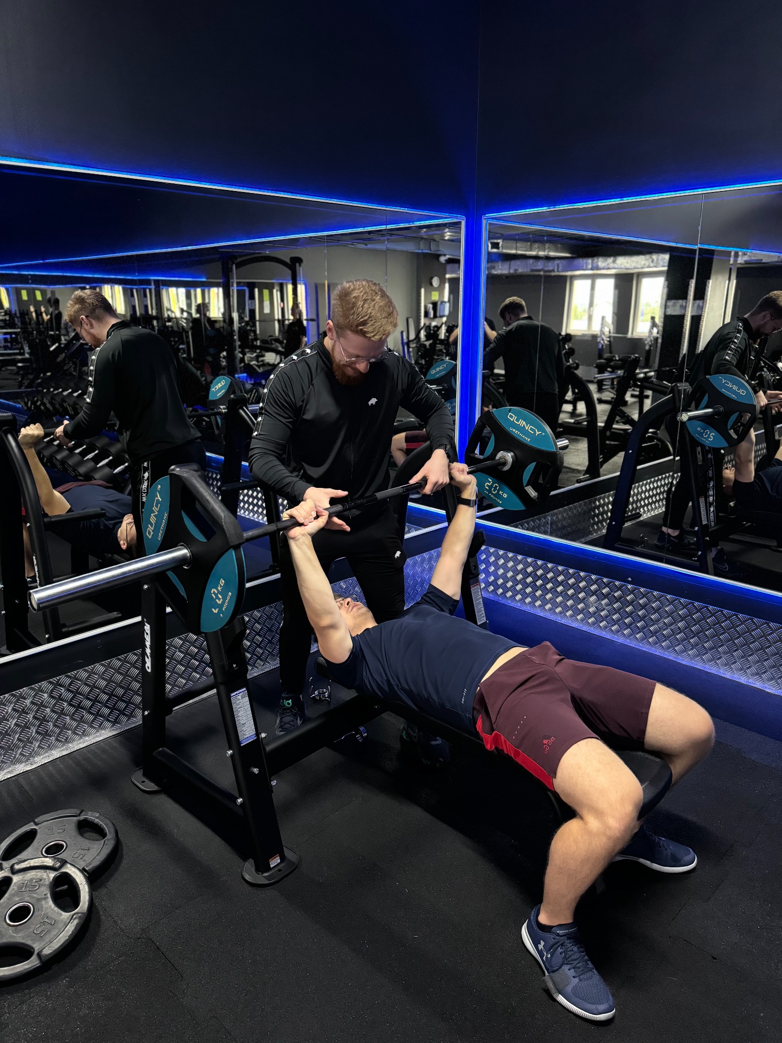 Mężczyzna ćwiczy wyciskanie sztangi na ławce pod nadzorem instruktora w nowoczesnej siłowni z lustrami i niebieskim oświetleniem LED.
