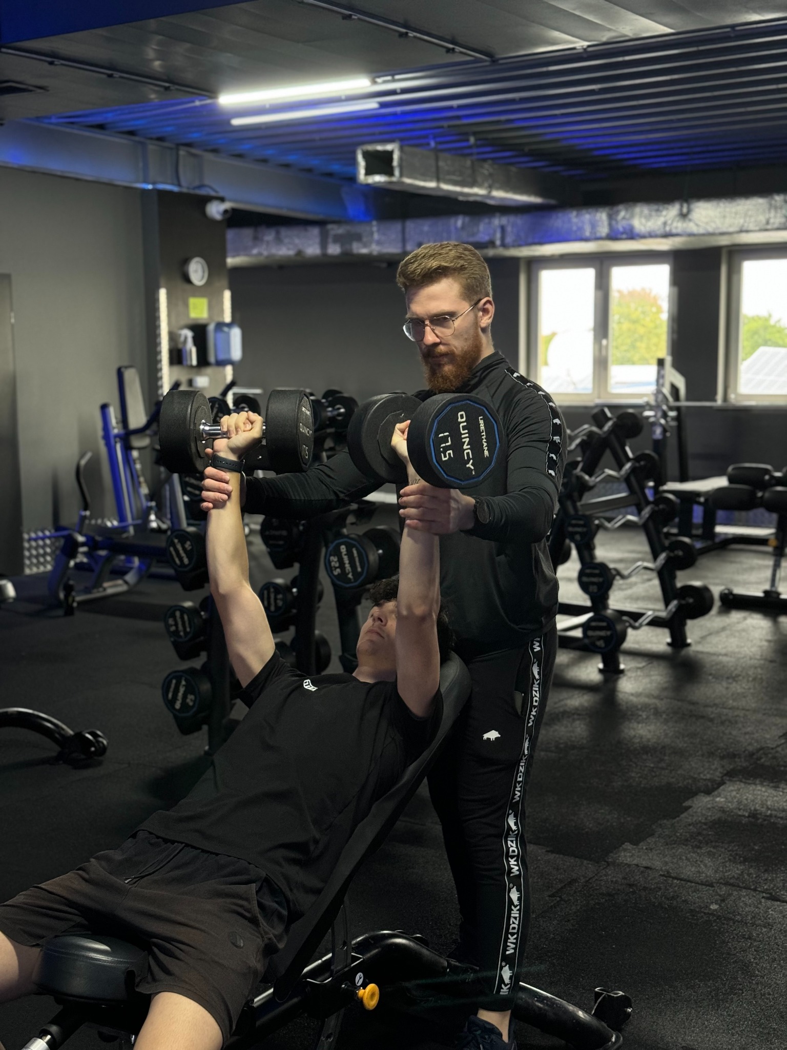 Mężczyzna z brodą w okularach asystuje mężczyźnie leżącemu na ławce podczas podnoszenia hantli w siłowni.