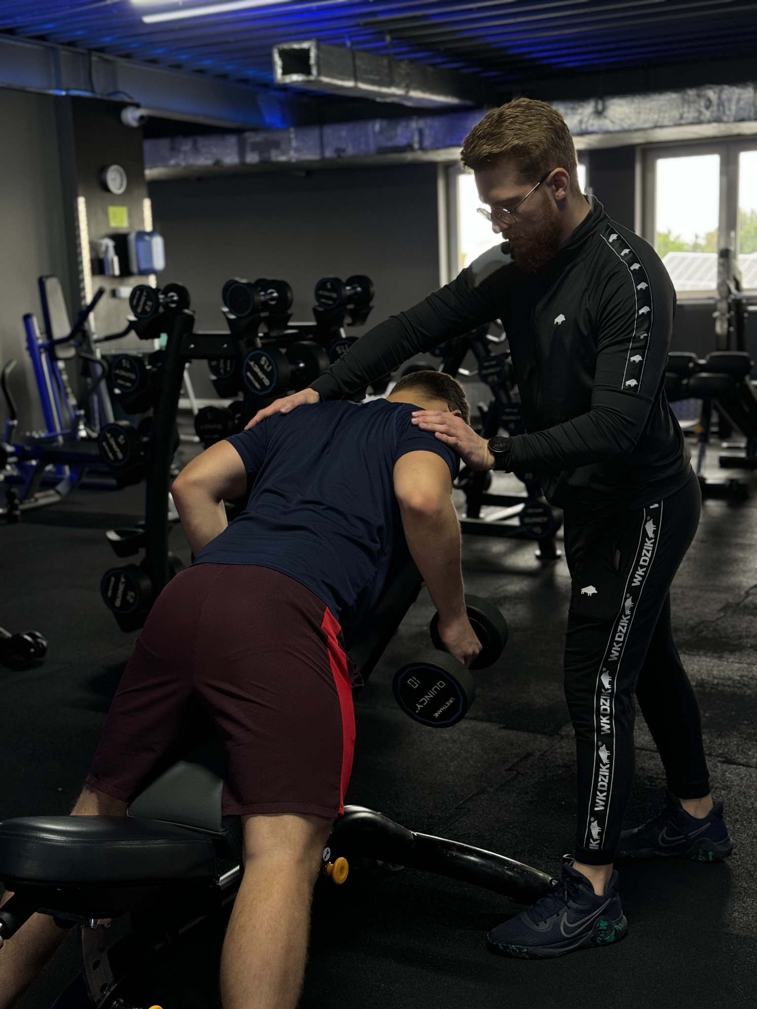 Trener personalny w czarnym dresie WK DZIK z ręką na plecach klienta ćwiczącego z hantlą na ławce w siłowni.