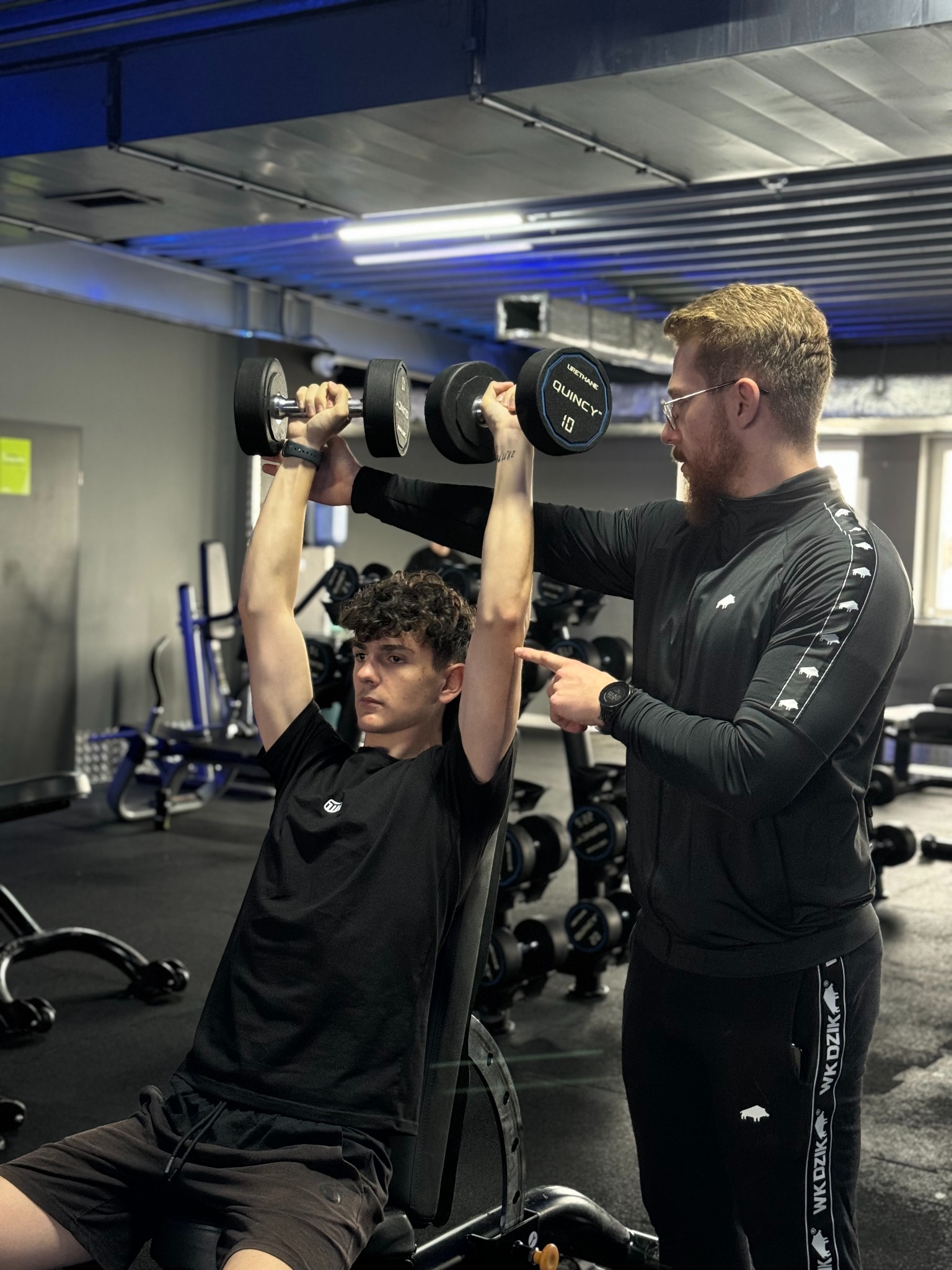 Młody mężczyzna ćwiczy podnoszenie hantli nad głową, instruowany przez trenera w czarnym dresie z białymi wzorami dzików, w tle siłownia z maszynami.