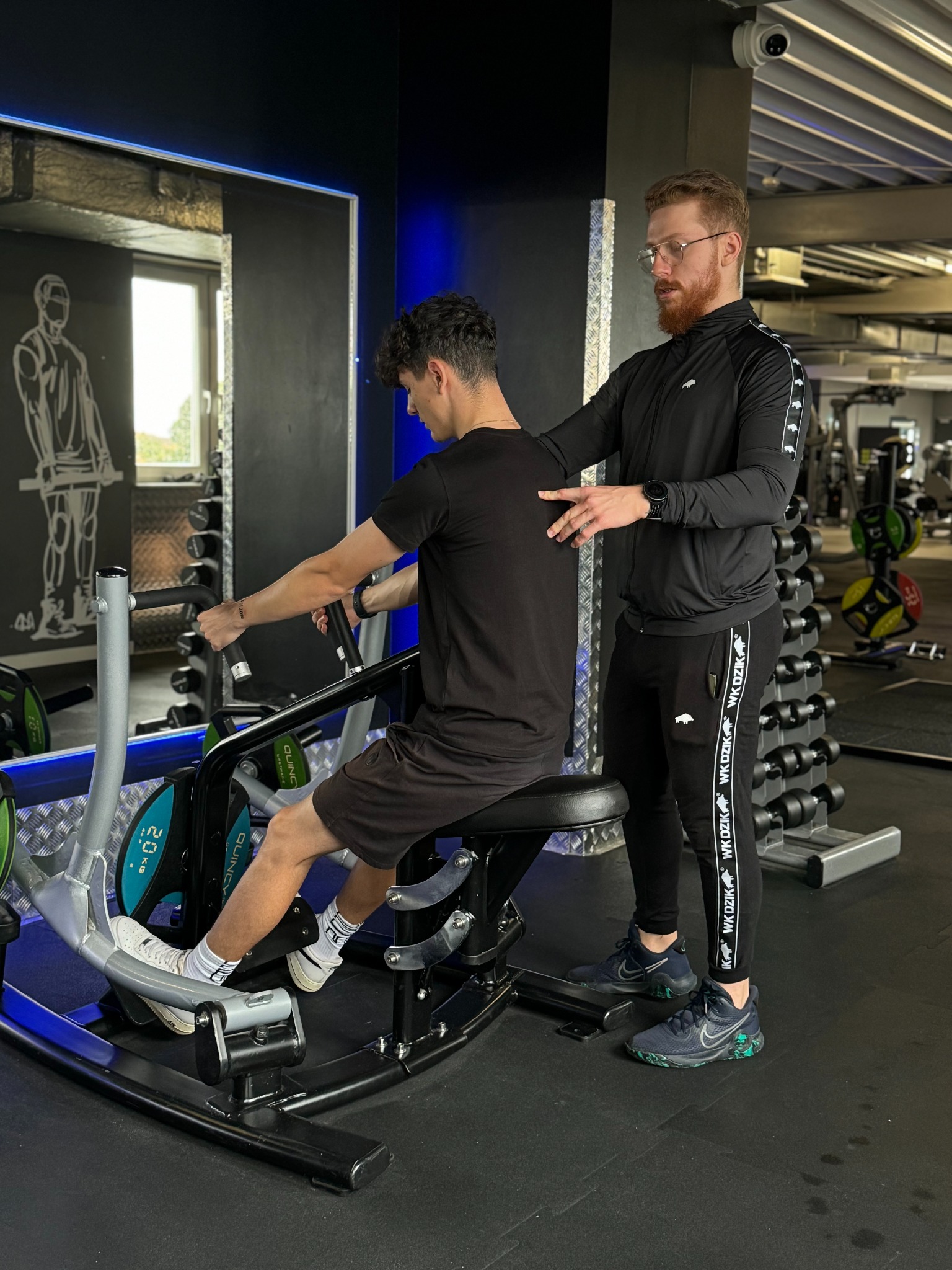 Młody mężczyzna ćwiczy na maszynie do wiosłowania w siłowni, nadzorowany przez trenera w czarnym dresie z logo WK DZIK.