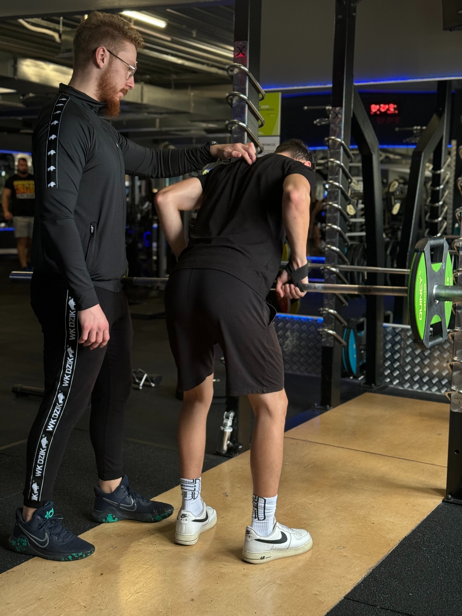 Mężczyzna w czarnej bluzie i spodniach z logo WK DZIK koryguje postawę mężczyzny przygotowującego się do podnoszenia ciężarów na siłowni.