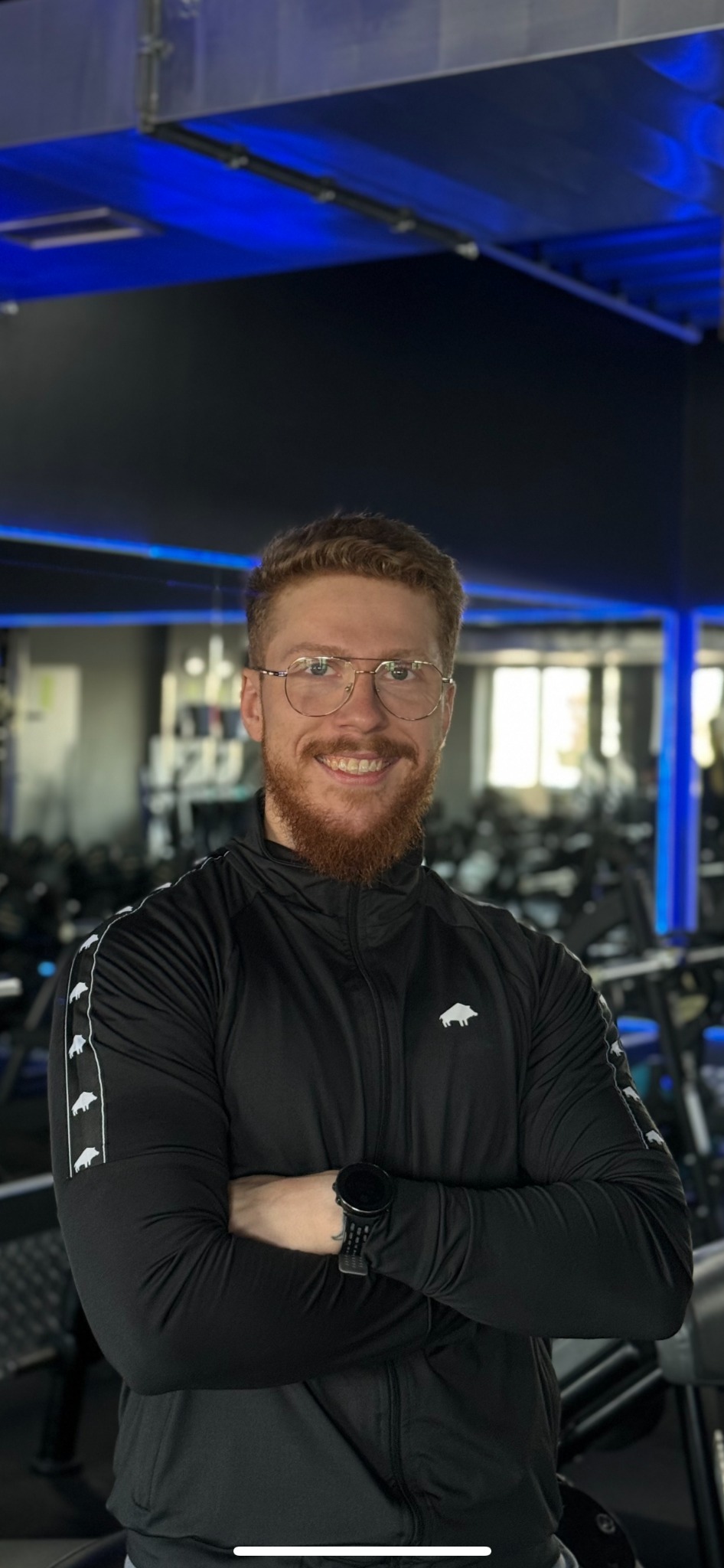 Uśmiechnięty mężczyzna z rudą brodą i okularach, ubrany w czarną bluzę sportową z logo dzika, stoi na tle siłowni z niebieskim oświetleniem LED