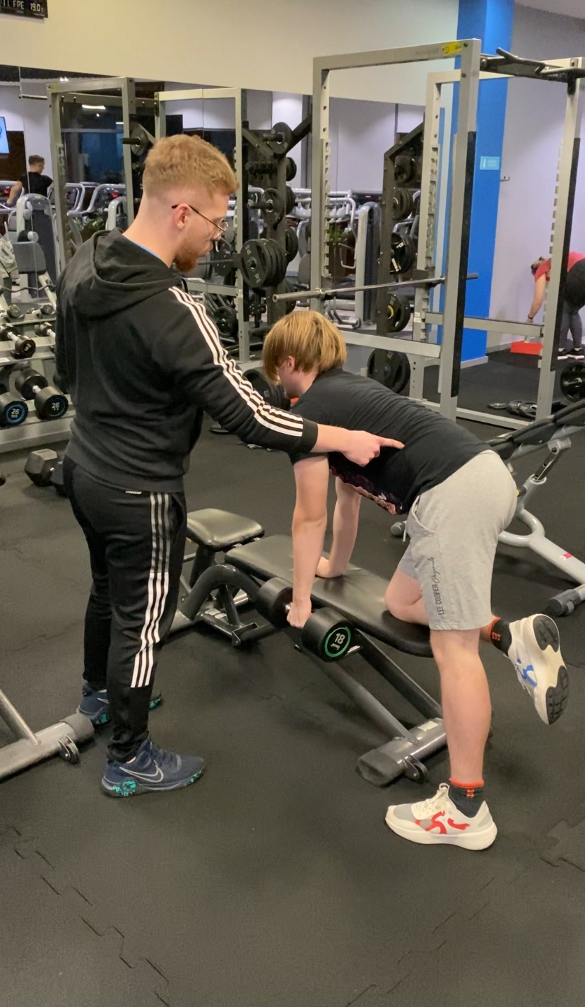 Trener personalny koryguje postawę klienta podczas ćwiczenia z hantlą na ławce w siłowni, widoczne ciężary i sprzęt do ćwiczeń w tle.