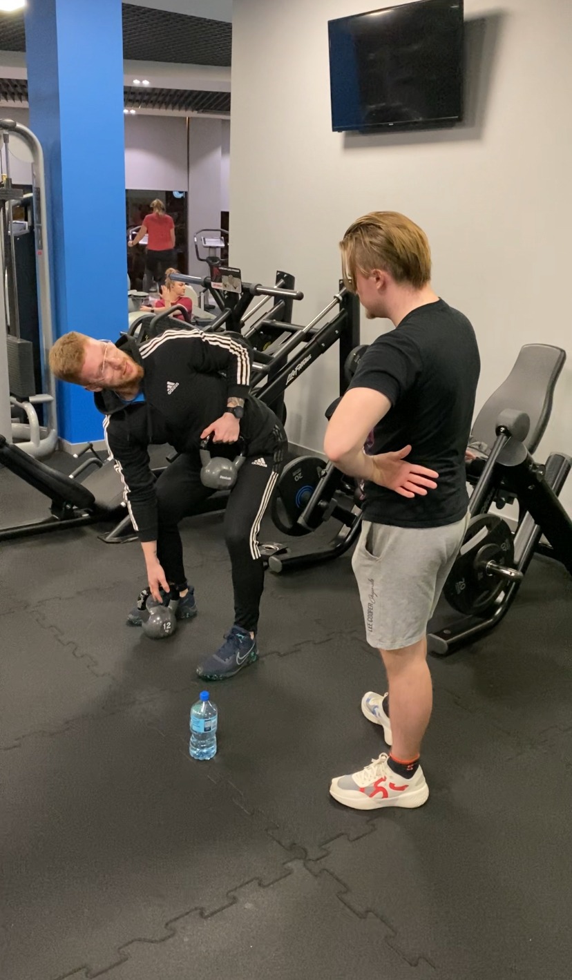 Mężczyzna w czarnym dresie Adidas wykonuje ćwiczenie z odważnikiem kettlebell w siłowni, nadzorowany przez drugiego mężczyznę w szarych spodenkach. Na podłodze butelka wody.