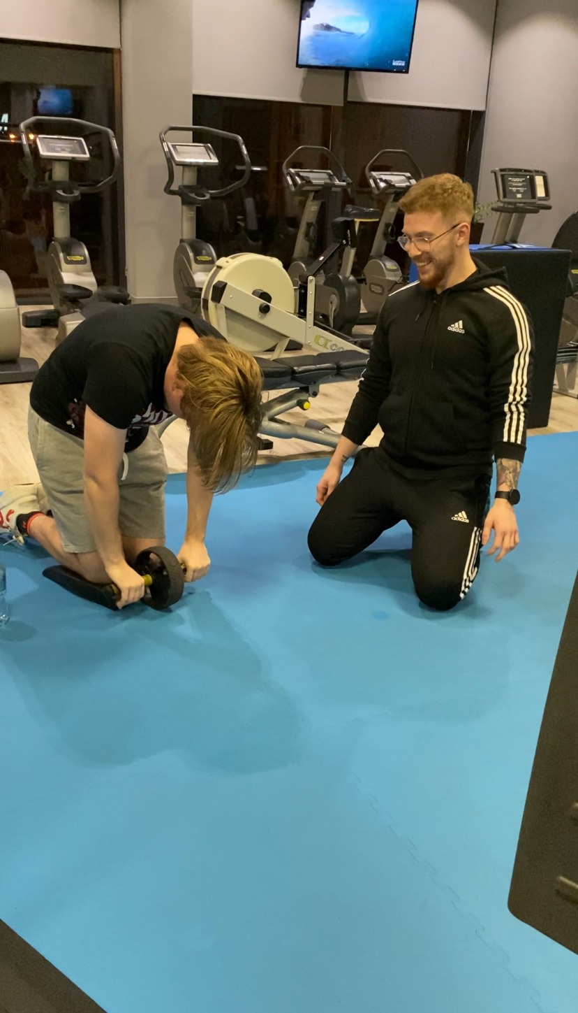 Mężczyzna ćwiczy na macie z kółkiem do ćwiczeń pod okiem trenera w czarnym dresie Adidas, w tle sprzęt do ćwiczeń i telewizor.