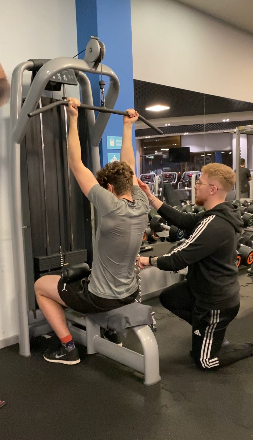 Mężczyzna ćwiczy na maszynie wyciąg górny pod nadzorem drugiego mężczyzny w sportowym stroju w siłowni. Widoczny tatuaż na ręce nadzorującego.