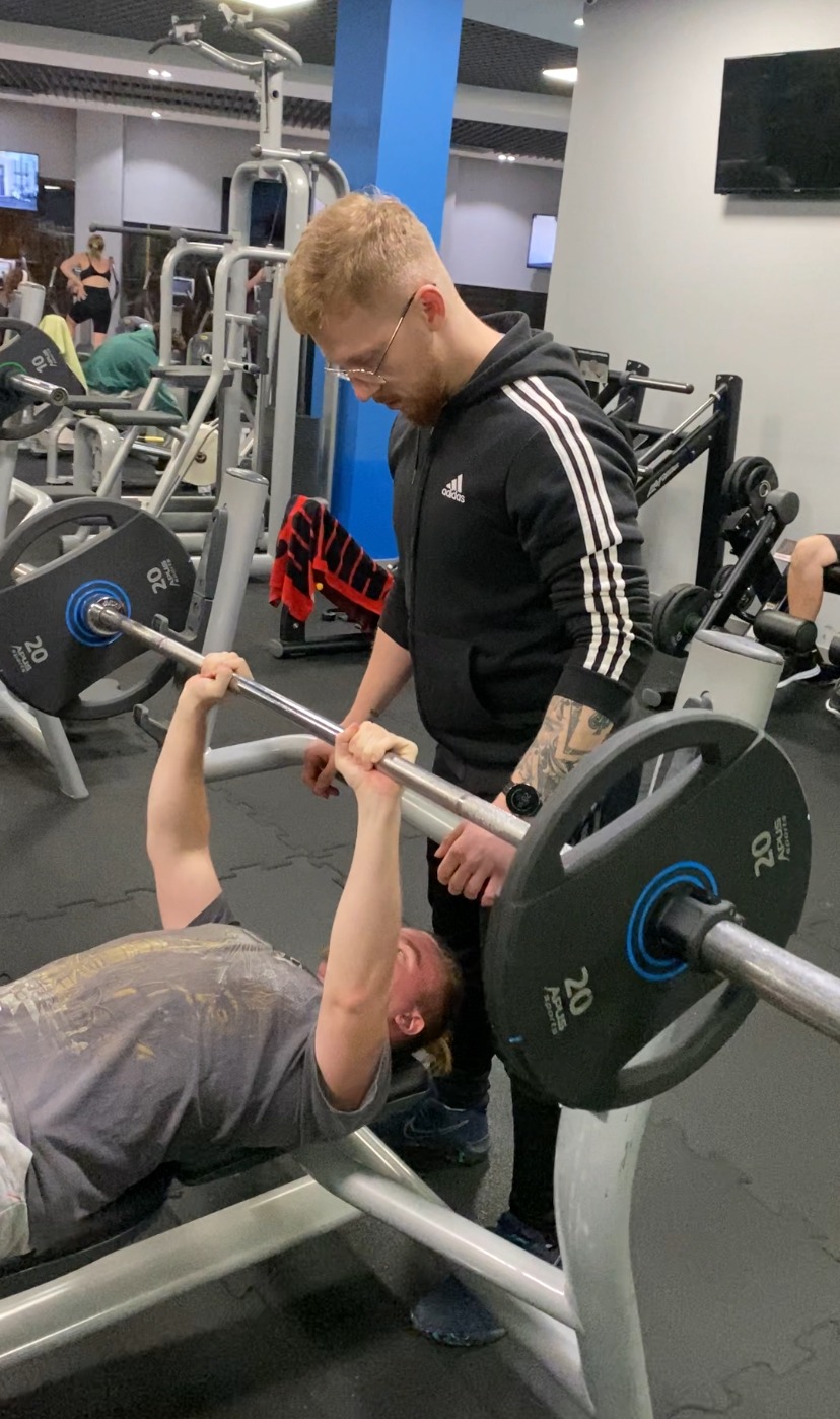 Mężczyzna w okularach, ubrany w czarną bluzę Adidas z trzema paskami, asekuruje ćwiczącego na ławeczce, który podnosi sztangę w siłowni.