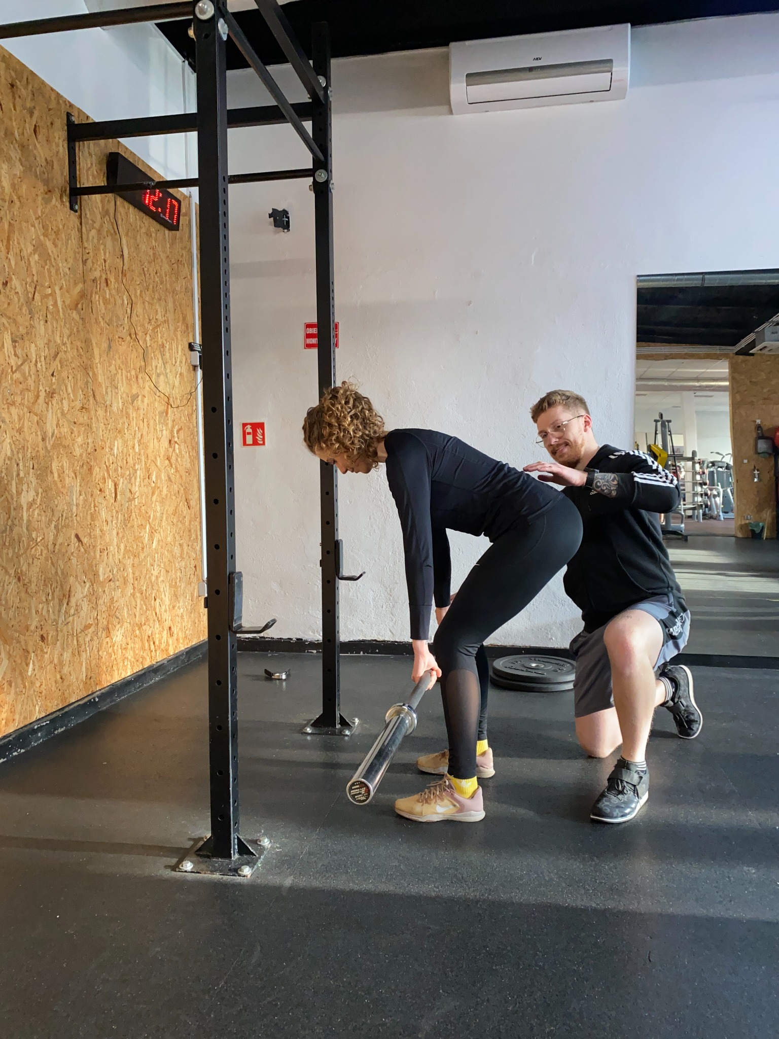 Kobieta w czarnym stroju podnosi sztangę pod nadzorem mężczyzny w okularach i sportowej bluzie, w tle ściana z zegarem w siłowni.