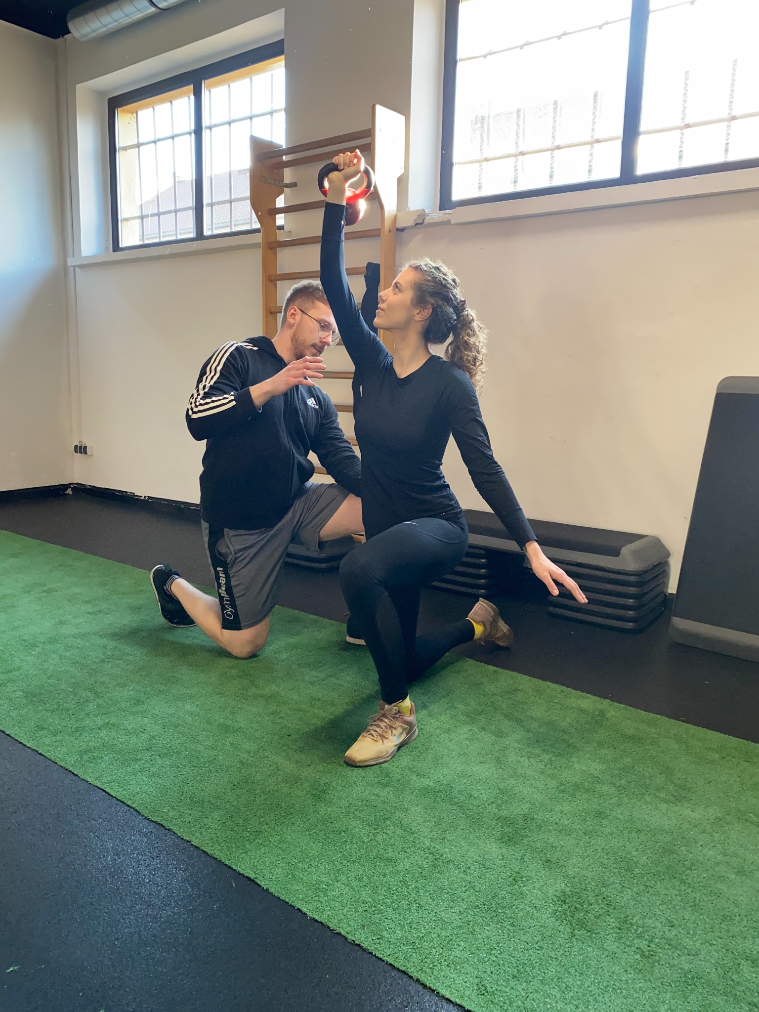 Kobieta ćwiczy z kettlebell pod nadzorem trenera personalnego w jasnej siłowni z drabinkami gimnastycznymi w tle.