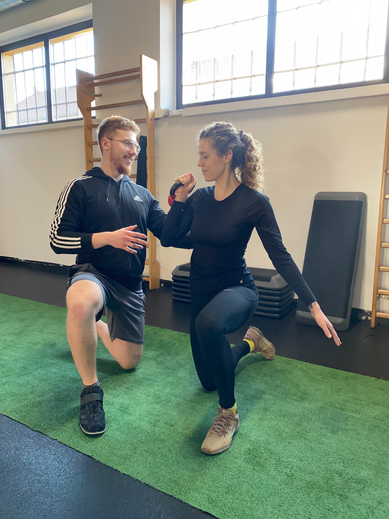 Kobieta ćwiczy z kettlebell pod okiem mężczyzny w bluzie Adidas, oboje w pozycji klęczącej na zielonej sztucznej trawie w siłowni.