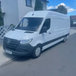 Biały samochód dostawczy Mercedes-Benz Sprinter zaparkowany na ulicy przed domem jednorodzinnym w słoneczny dzień.