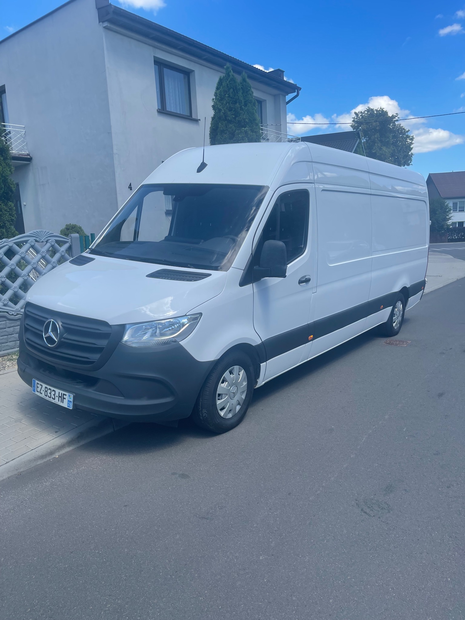 Biały samochód dostawczy Mercedes-Benz Sprinter zaparkowany na ulicy przed domem jednorodzinnym w słoneczny dzień.