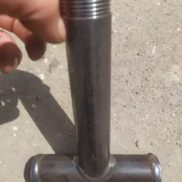 Metalowa rura w kształcie litery T z widocznymi spawami, leżąca na betonowym podłożu, z fragmentem dłoni w kadrze.