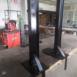 Dwie czarne, metalowe kolumny konstrukcyjne z podstawami, świeżo pomalowane, stojące w warsztacie, widoczny sprzęt do spawania i butla gazowa w tle.