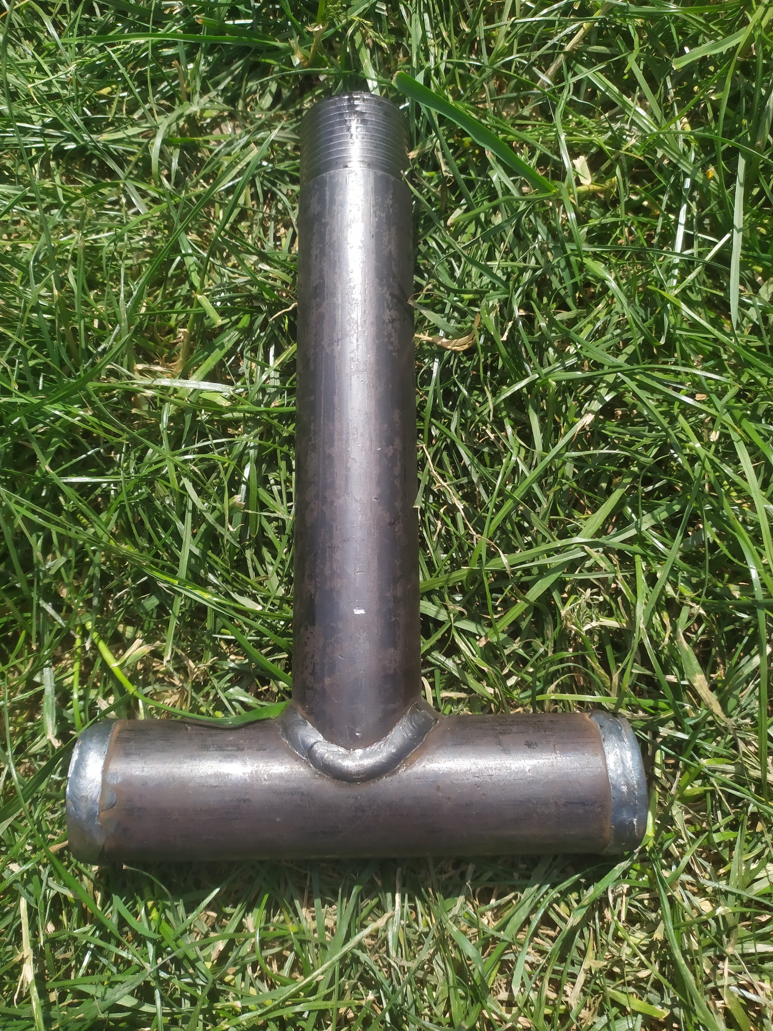 Metalowa rura w kształcie litery T z widocznymi śladami spawania, leżąca na trawie w jasnym świetle słonecznym.