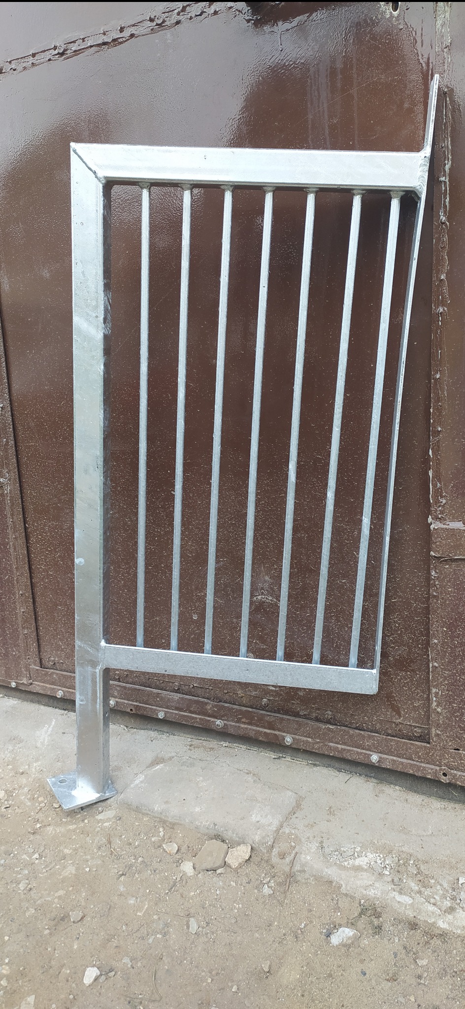 Metalowa barierka ochronna z pionowymi prętami, przyspawana do ramy, ocynkowana, widoczna na tle brązowych drzwi.