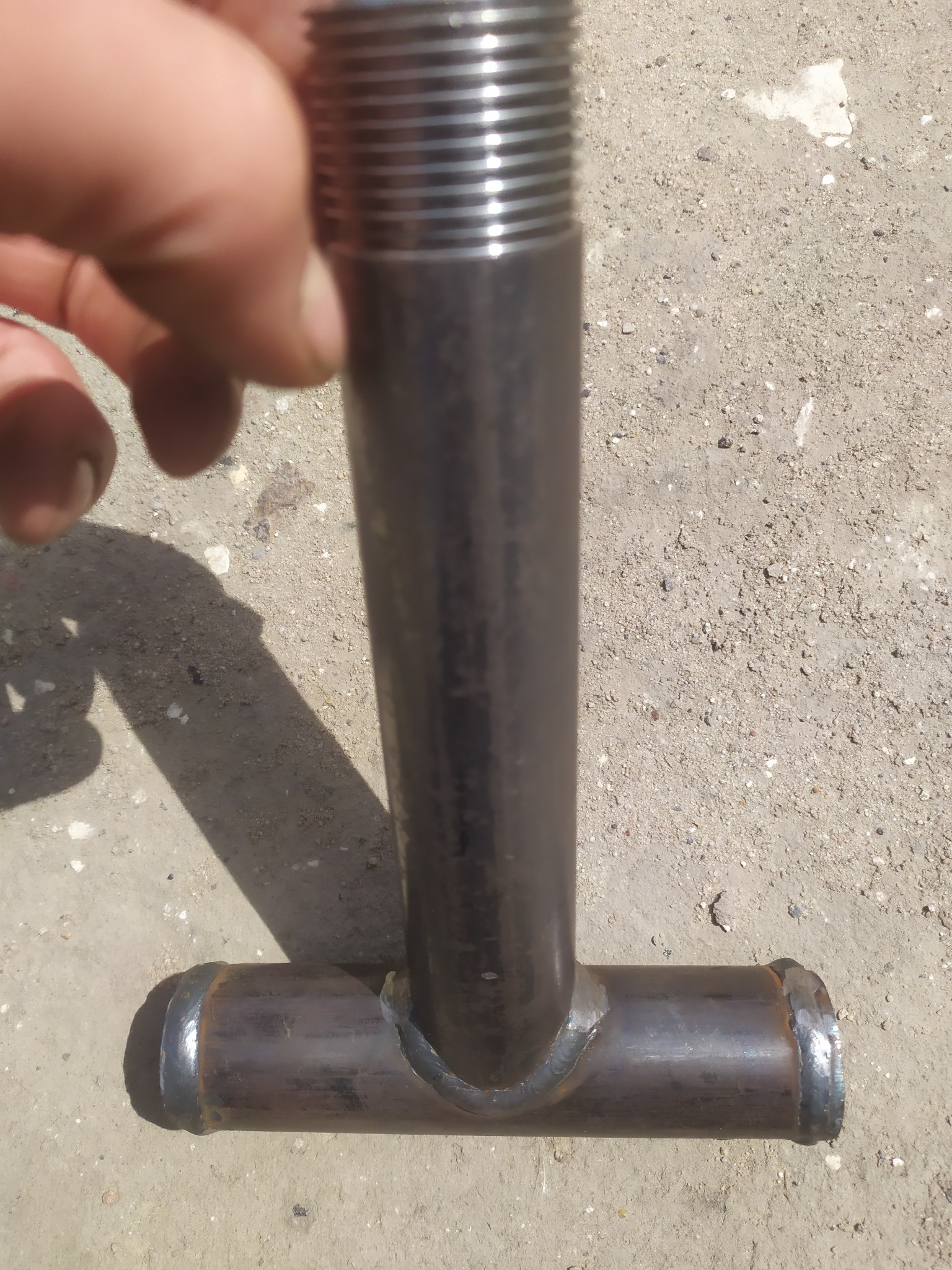 Metalowa rura w kształcie litery T z widocznymi spawami, leżąca na betonowym podłożu, z fragmentem dłoni w kadrze.