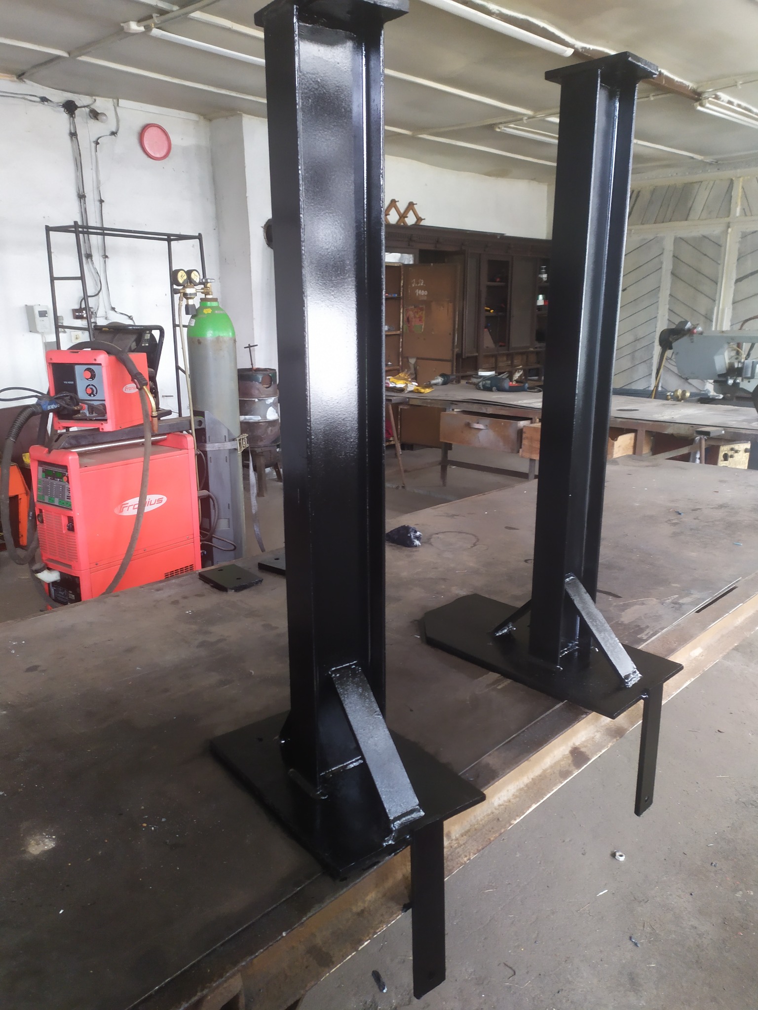 Dwie czarne, metalowe kolumny konstrukcyjne z podstawami, świeżo pomalowane, stojące w warsztacie, widoczny sprzęt do spawania i butla gazowa w tle.