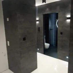 Nowoczesna łazienka z ciemnoszarymi płytkami o fakturze betonu, białymi podłogami, minimalistycznym oświetleniem punktowym i wiszącą toaletą w głębi.