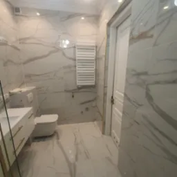 Elegancka łazienka wykończona w marmurkowych płytkach, z białą armaturą i złotymi akcentami na szafce umywalkowej oraz obramowaniu drzwi.