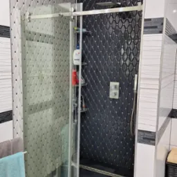 Nowoczesna kabina prysznicowa z przesuwnymi drzwiami, wyłożona czarno-białymi płytkami w romby, z półkami na kosmetyki i odpływem liniowym.