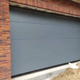 Zamknięta, szara, segmentowa brama garażowa z poziomymi przetłoczeniami, wkomponowana w ceglaną elewację budynku w trakcie budowy, widoczne pozostałości gruzu i piasku.