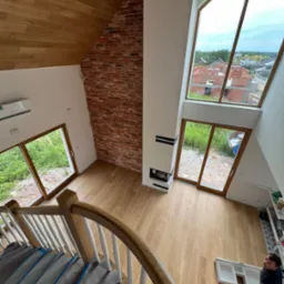 Wysoki salon z drewnianą podłogą i schodami zabezpieczonymi folią, ceglana ściana, duże okna z widokiem na budowę, montażysta mebli w rogu.