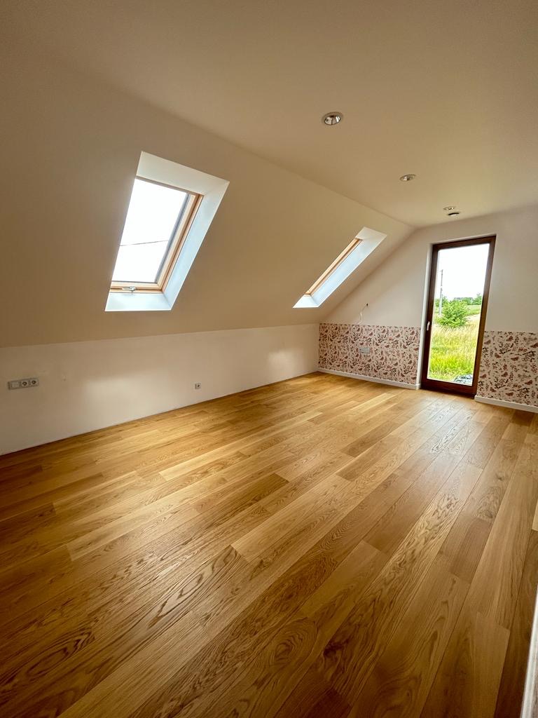 Jasne, przestronne poddasze z drewnianą podłogą, dwoma oknami dachowymi i wzorzystą tapetą na ścianie przy drzwiach balkonowych.