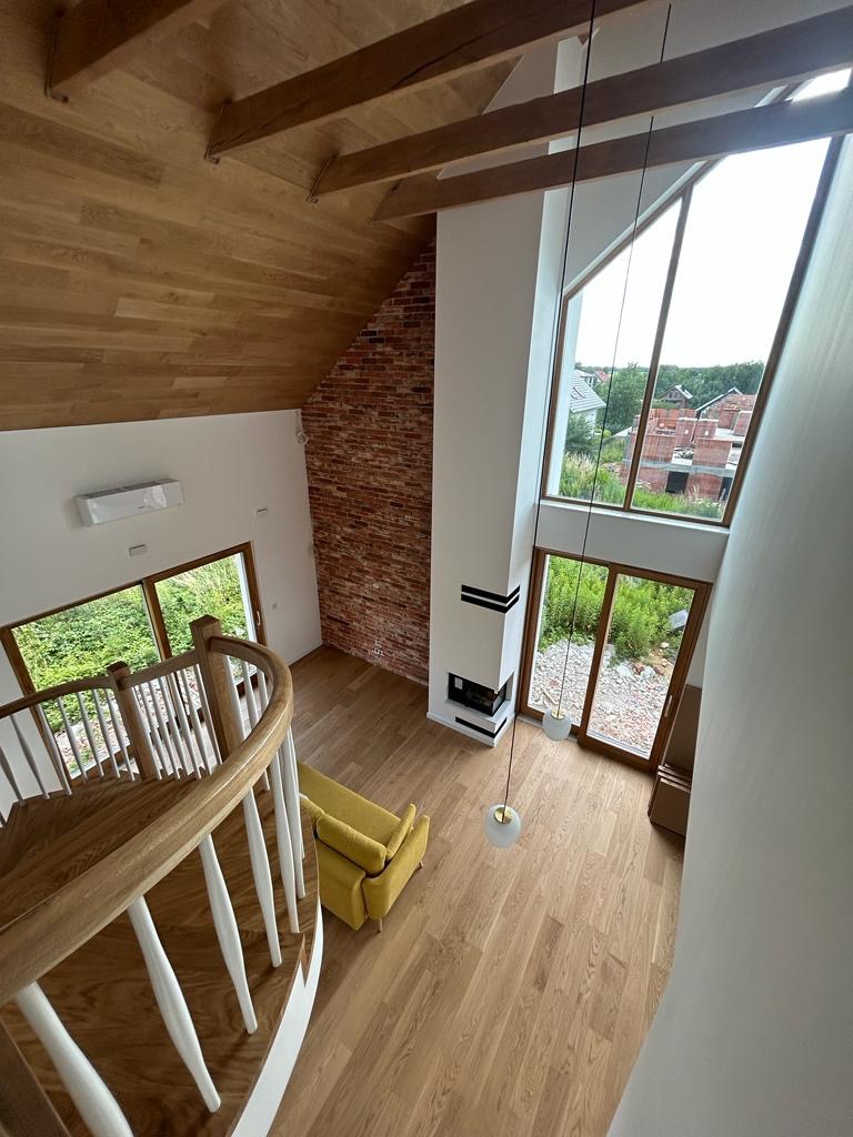 Wysoki salon z drewnianą podłogą, ceglaną ścianą, spiralnymi schodami, dużymi oknami i minimalistycznym kominkiem, widziany z góry.