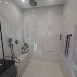 Nowoczesna kabina prysznicowa z deszczownicą, wykończona białymi płytkami imitującymi marmur z czarnymi żyłami, czarna armatura i szklana ściana.