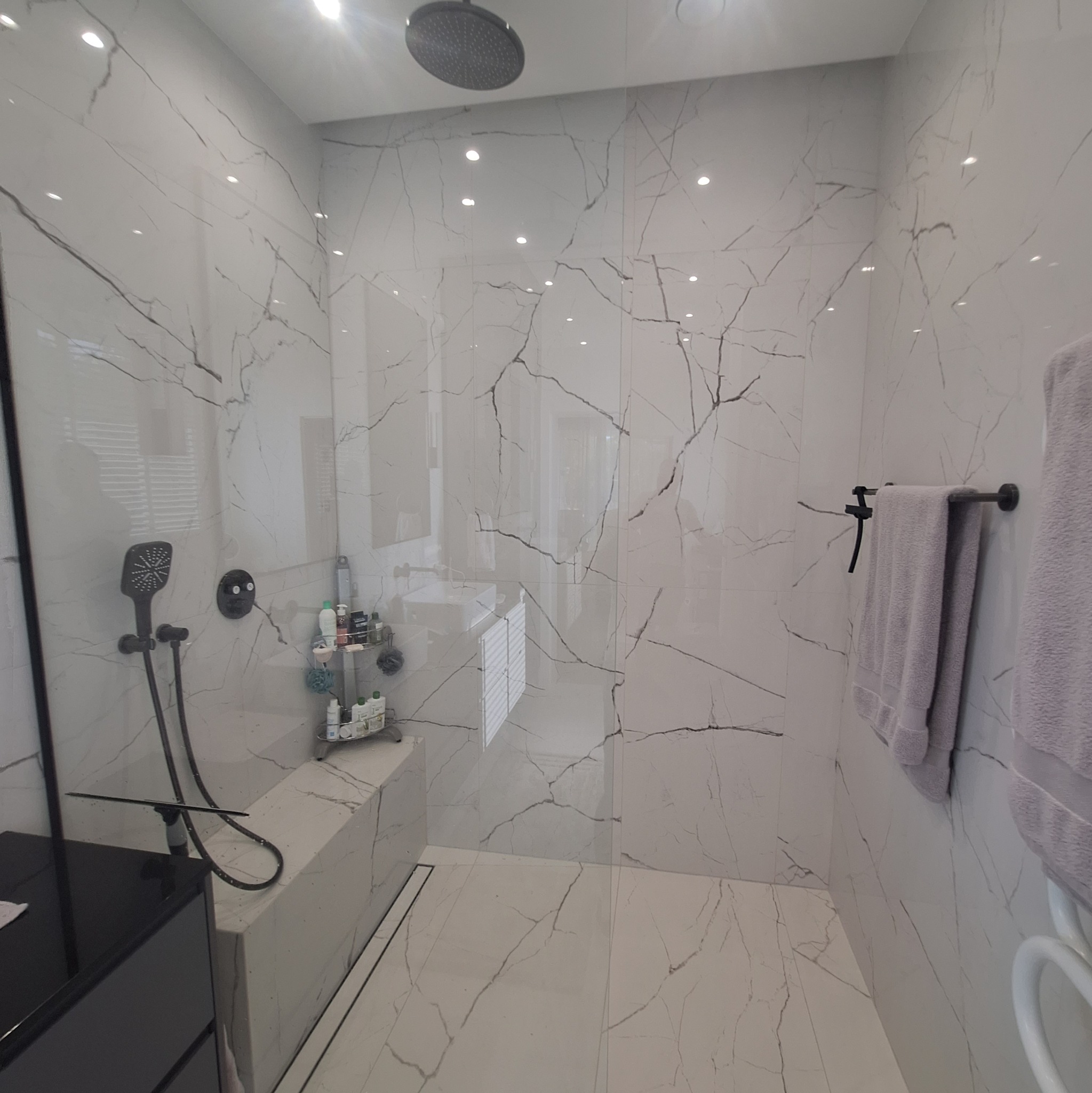 Nowoczesna kabina prysznicowa z deszczownicą, wykończona białymi płytkami imitującymi marmur z czarnymi żyłami, czarna armatura i szklana ściana.