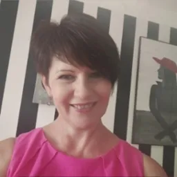 Portret uśmiechniętej kobiety z krótkimi brązowymi włosami, w różowej bluzce, na tle ściany w czarno-białe pasy i obrazu z sylwetką kobiety w czerwonym kapeluszu.