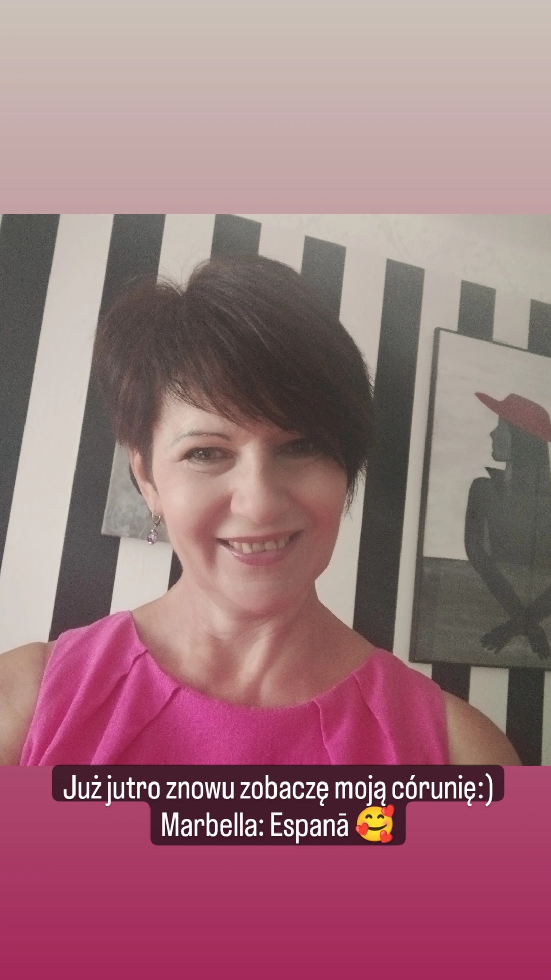 Portret uśmiechniętej kobiety z krótkimi brązowymi włosami, w różowej bluzce, na tle ściany w czarno-białe pasy i obrazu z sylwetką kobiety w czerwonym kapeluszu.