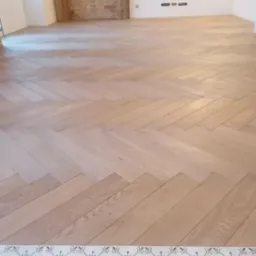 Jasny parkiet ułożony we wzór jodełki francuskiej, z dekoracyjnym pasem płytek ceramicznych przy ścianie, na tle surowej ściany z cegły.