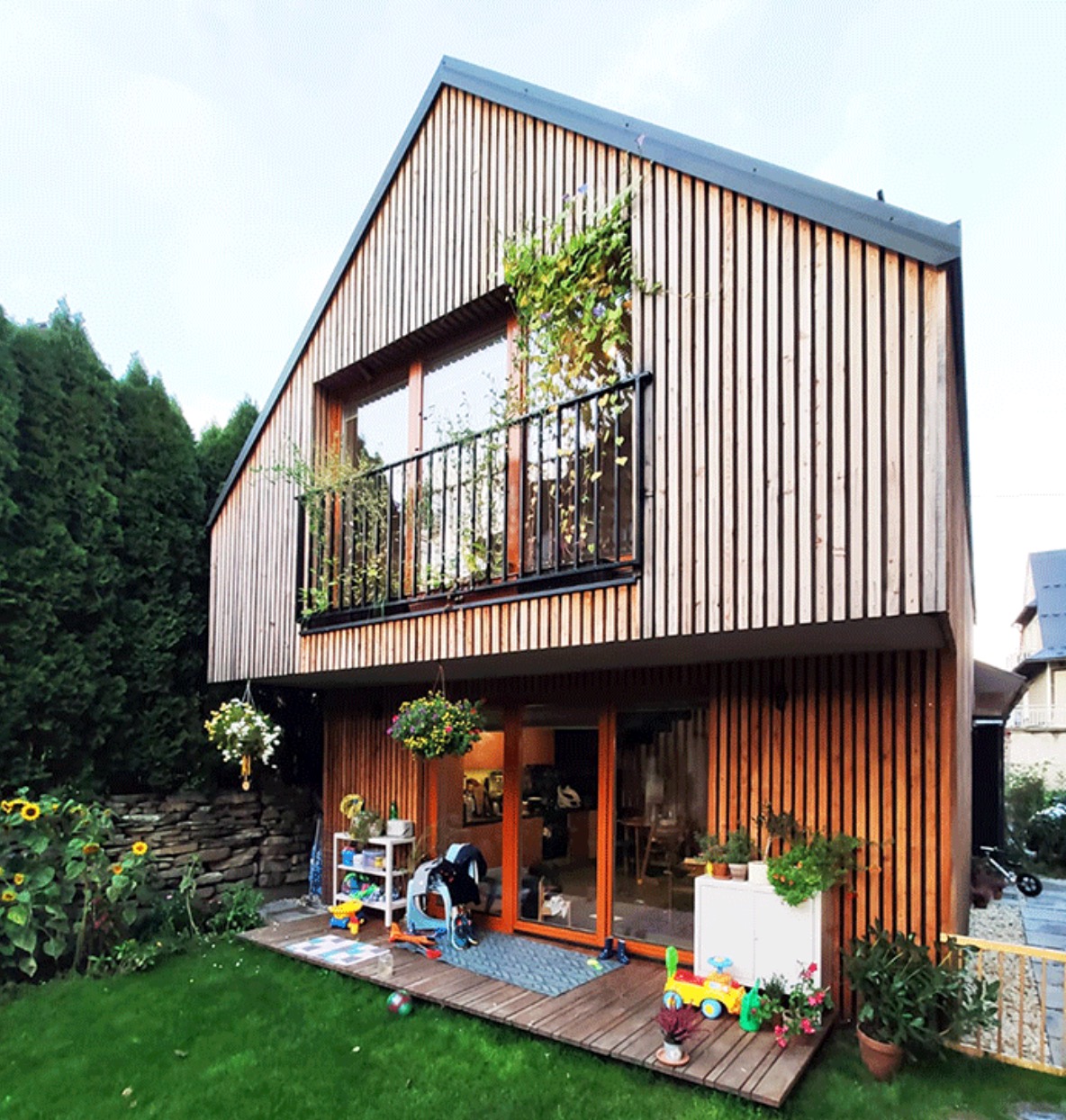 Dwukondygnacyjny dom z drewnianą elewacją wertykalną, balkonem z roślinami i tarasem z zabawkami dla dzieci.
