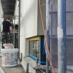 Pracownik na rusztowaniu wyrównuje świeżo nałożoną warstwę gładzi na elewacji budynku, widoczny fragment okna zabezpieczonego folią i taśmą malarską, obok stoi wiadro z masą szpachlową.