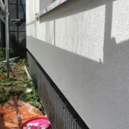 Świeżo otynkowana elewacja budynku w Paczkowie, z widocznym oknem zabezpieczonym folią i fragmentem rusztowania, teren wok&oacute;ł budynku z pozostałościami po pracach budowlanych.