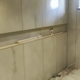 Ściana z marmurowymi płytkami w trakcie wykańczania, widoczne przewody i pozostałości po kleju na podłodze, zabezpieczonej folią.