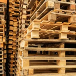 Stos drewnianych palet ułożonych równo jeden na drugim, widoczne stemple i oznaczenia na niektórych paletach.