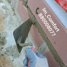 Murowanie ściany z cegły klinkierowej, widoczna kielnia z zaprawą w dłoni w rękawicy oraz poziomica na górze muru.